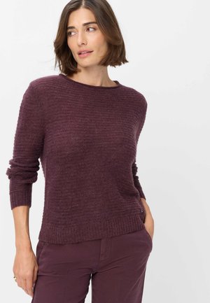 Donna con capelli corti castani che indossa un maglione viola scuro testurizzato e pantaloni abbinati, che guarda leggermente verso sinistra su uno sfondo bianco.