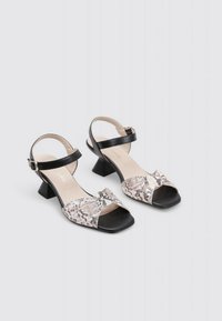Sandalias de tacón bloque de cuero negro con diseño entrelazado que presenta un upper textil brillante en plata y beige. Punta cuadrada, correa ajustable.