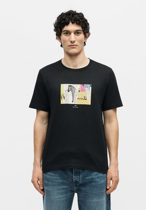 Άνδρας που φοράει μαύρο μπλουζάκι με πολύχρωμο ορθογώνιο γραφικό μιας ζέβρας και αφηρημένων σχημάτων, σε συνδυασμό με μπλε τζιν, πάνω σε λευκό φόντο.