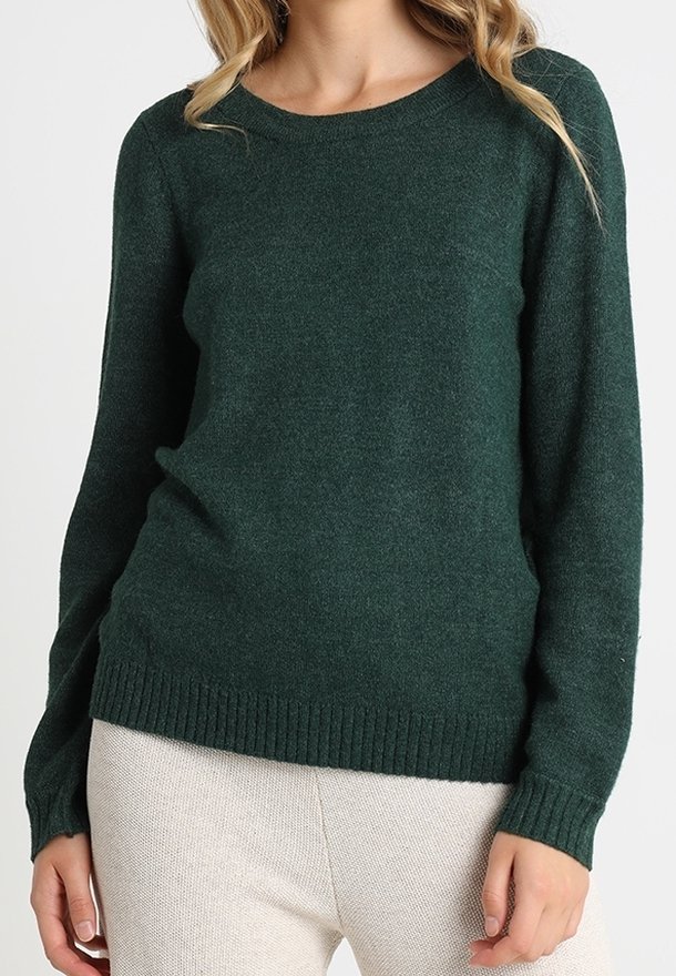 Femme portant un pull en maille vert foncé à manches longues avec des poignets et un ourlet côtelés, associé à un pantalon clair à texture.