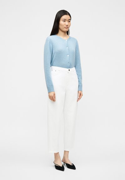 Γυναίκα που φοράει ανοιχτό μπλε πουλόβερ με κουμπιά, ψηλόμεσο λευκό παντελόνι και μαύρες μυτερές γόβες με λουράκι πίσω, στέκεται μπροστά σε απλό φόντο.