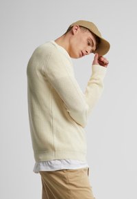Suéter de punto color crema con textura acanalada y mangas largas, combinado con una gorra de béisbol beige y una camisa clara debajo.