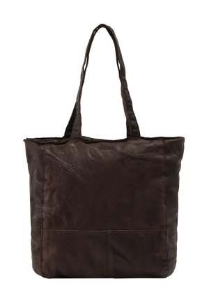Donna con capelli lunghi castani che indossa un maglione beige e pantaloni bianchi, porta una grande borsa tote in pelle marrone scuro sulla spalla.