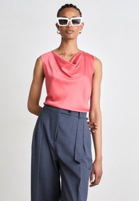 Σατέν χωρίς μανίκια μπλουζάκι σε κοραλί απόχρωση με δροσερό ντεκολτέ, συνδυασμένο με ψηλόμεσα, πλισέ γκρι παντελόνια. Λευκά τετράγωνα γυαλιά ηλίου ολοκληρώνουν την εμφάνιση.