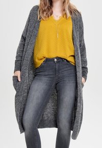Vrouw met mosterdgele blouse, lange grijze cardigan, donkergrijze skinny jeans, met handen in de zakken van de cardigan, staand tegen een effen achtergrond.