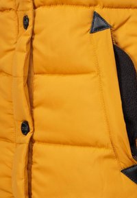 Yellow padded jacket with black buttons, featuring a textured pocket and a smooth finish, highlighting quilted detailing and contrasting accents.