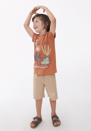 Camiseta de manga corta marrón con gráficos de cactus y texto, combinada con pantalones cortos beige. El niño lleva sandalias oscuras, sonriendo con los brazos levantados.