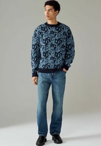 Tengerés kék paisley mintás kötött pulóver kerek nyakkal, világoskék farmernadrággal és fekete cipővel párosítva. Texturált anyag és laza szabás.