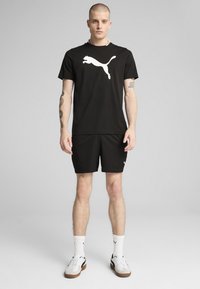Musta urheilupaita, jossa on suuri valkoinen Puma-logo, yhdistettynä mustiin shortseihin ja valkoisiin sukkiin. Mallilla on päällään valkoiset ja ruskeat urheilujalkineet.