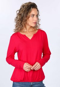 Rotes langärmliges Oberteil mit V-Ausschnitt, gerafftem Bund und weicher Textur. Modell trägt das Shirt, Hände vor dem Körper, mit lässigen Jeans.