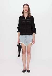 Črna bluzica s plastmi in čipkastimi rokavi, v kombinaciji z svetlim jeans krilom z raztrganimi robovi. Model drži teksturirano črno torbico.