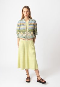 Daugiaspalvė trumpa bomber tipo striukė su gėlių ir geometriniais raštais, dėvima ant geltonos plišinės maksi suknelės, derinama su rudomis šlepetėmis ant dirželio.