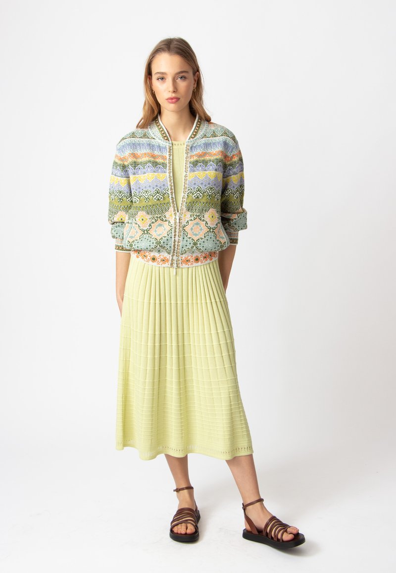 Daugiaspalvė trumpa bomber tipo striukė su gėlių ir geometriniais raštais, dėvima ant geltonos plišinės maksi suknelės, derinama su rudomis šlepetėmis ant dirželio.