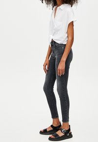 Jeans ajustados grises de mezclilla, combinados con una camiseta blanca de manga corta atada a la cintura y sandalias negras de tiras con un logo.