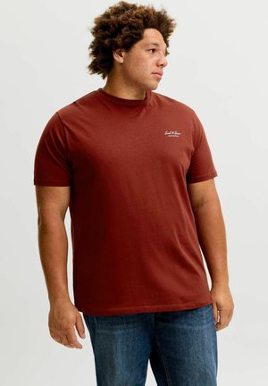 Homme aux cheveux bouclés portant un t-shirt rouge rouille et un jean bleu, regardant vers la droite sur un fond clair uni.