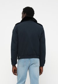 Navy blauwe gewatteerde jas met een faux fur kraag en schouderepauletten. Voorzien van knoopmanchetten en een getailleerde taille. Gezien vanaf de achterkant.