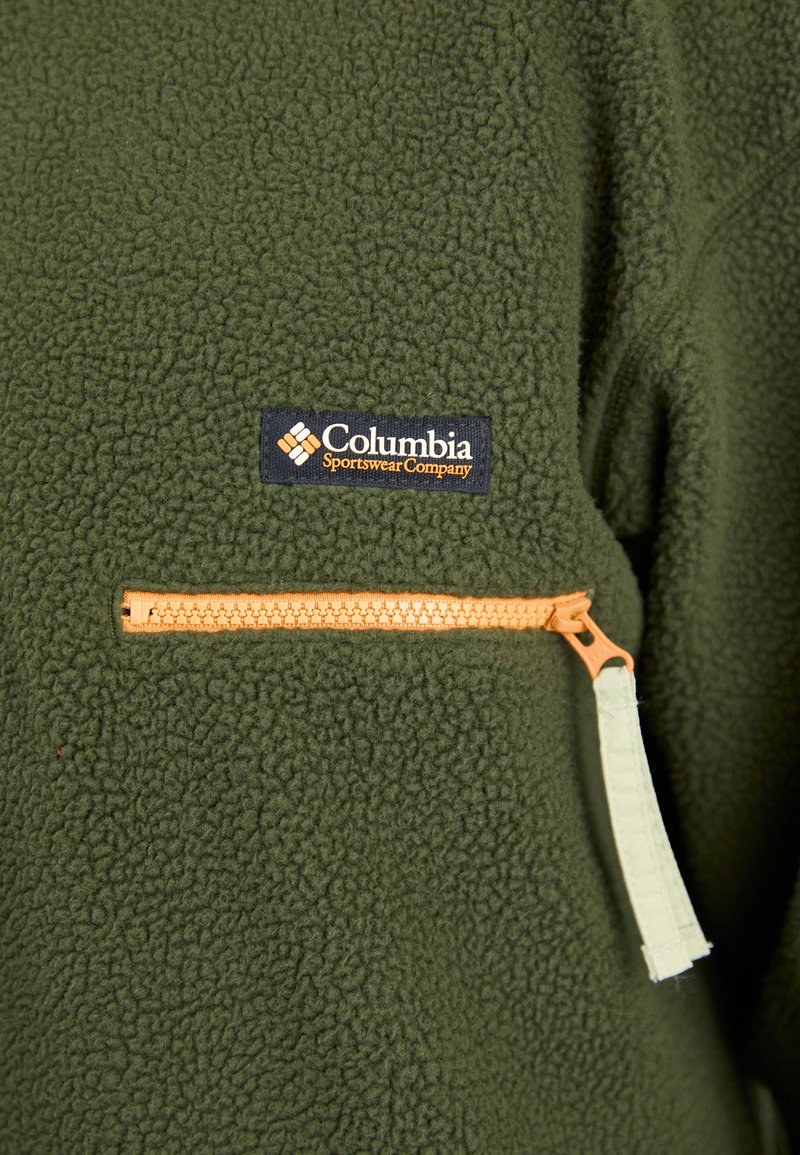 Olijfgroene fleecejack met een gestructureerd oppervlak, een met ritssluiting afgesloten zak met een oranje ritssluiting, en een marineblauw logo-embleem van Columbia Sportswear.