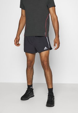 Schwarze Sportshorts und ein dunkelgraues, strukturiertes T-Shirt mit roten Akzenten. Die Shorts haben weiße Streifen und ein Adidas-Logo. Schwarze Sneaker runden das Outfit ab.