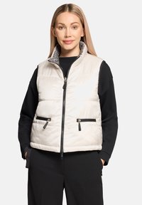 Kremowa pikowana kamizelka z czarnym zamkiem błyskawicznym, bocznymi kieszeniami oraz wzorzystym, wewnętrznym kołnierzem. Noszona na czarnej bluzce z długim rękawem.