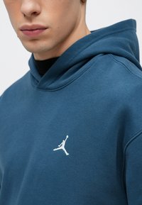 Φούτερ με κουκούλα, ύφασμα σε τιρκουάζ απόχρωση, μαλακή υφή, με λευκό λογότυπο Jumpman στο στήθος. Κοντινό πλάνο στον λαιμό και το πρόσωπο.