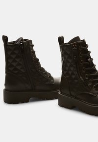 Botas negras hasta el tobillo con textura acolchada, cordones en la parte delantera, ojales de metal y cremallera lateral. Suela de goma gruesa con un ligero tacón.