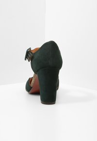 Chaussure à talons hauts en daim vert foncé avec un dos arrondi, une bride à la cheville et des accents texturés dorés sur les côtés. Semelle brune visible.
