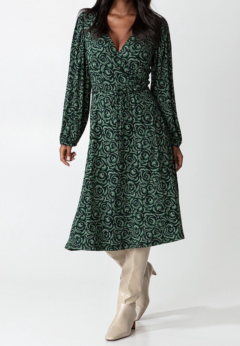 Femme portant une robe portefeuille fleurie verte avec de longues manches bouffantes et des bottes beige montant jusqu'aux genoux, debout devant un fond blanc uni.