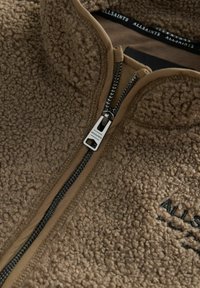 Brun fleece zip jakke med høj krave, tekstureret overflade, sort lynlås og broderet logo. Blødt materiale med holdbar syning.