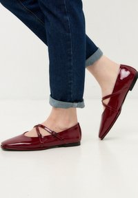 Bordeauxfarbene Lackleder-Ballerinas mit Kreuzriemen, abgerundeter Zehenpartie und niedrigem Absatz, kombiniert mit gerollten Jeanshosen. Glänzende Textur und elegantes Design.