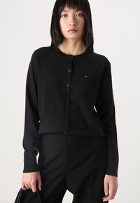 Zwarte gebreide cardigan met knopen, ronde halslijn en geribbelde manchetten. Bevat een klein gekleurd logo op de linkerkant van de borst. Gedirigeerd over zwarte broek.