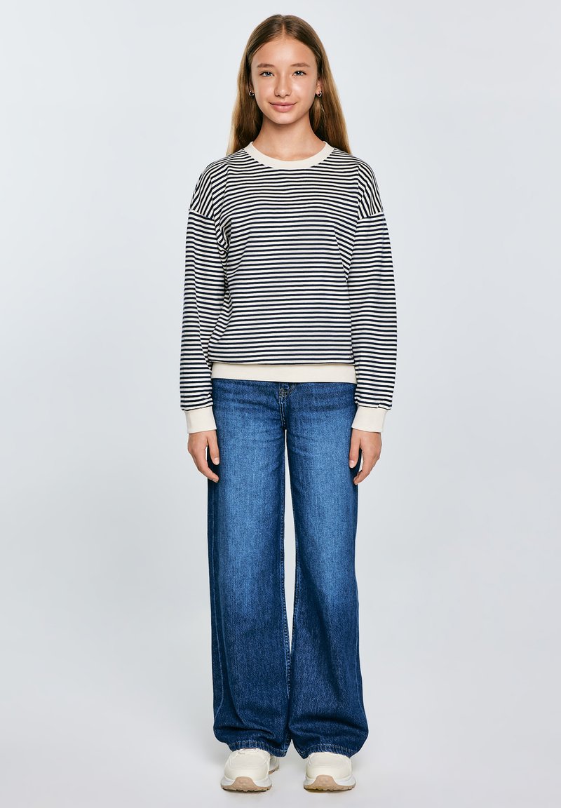 Dryžuotas džemperis baltai ir tamsiai mėlynai, apvaliu kaklu, laisvas fasonas, derinamas su plačiomis mėlynomis džinsinėmis kelnėmis ir baltais sportbačiais. Kasdienis apranga.