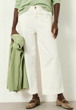 Weiße, weit geschnittene Hosen mit hohem Bund, kombiniert mit einem grün gerippten Pullover. Die Person hält den Pullover und trägt braune Wildlederschuhe.