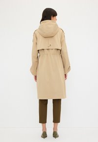 Beige trenchcoat med huva, cinchad midja och vida ärmar, i kombination med bruna beskurna byxor och gröna klackskor. Minimalistisk design.