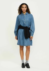Rochie camasa din denim de culoare albastru deschis, cu un guler tip buton, maneci lungi si un pulover tricotat negru legat la talie.
