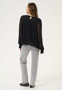 Blusa nera a maniche lunghe con design fluido, schiena arricciata e dettaglio a chiave nel collo, abbinata a jeans grigi chiari a gamba dritta.