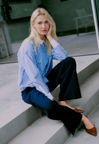 Blauw-wit gestreepte blouse met lange mouwen, gecombineerd met wijde marineblauwe broeken en bruine puntige schoenen, zittend op grijze treden.