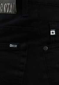 Nahaufnahme schwarzer Denim-Jeans mit Vordertasche, auf der ein "Big Star"-Etikett und ein kleines weißes Schild mit einem schwarzen Sternsymbol zu sehen sind.