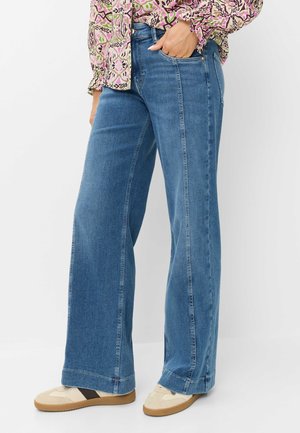 Kvinde iført blå vidde ben jeans, beige sneakers med brune striber og en mønstret langærmet bluse, hånd i lommen, stående på en hvid baggrund.