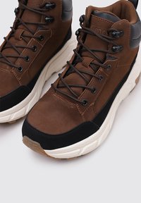 Zapatillas de medio corte sintéticas y de ante marrón con acentos en negro, suelas texturizadas y cordones negros con ojales de metal.