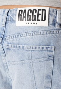 Jeans de mezclilla azul claro que presentan una etiqueta blanca con texto negro que dice "RAGGED JEANS", con costuras visibles y un detalle en el bolsillo trasero.