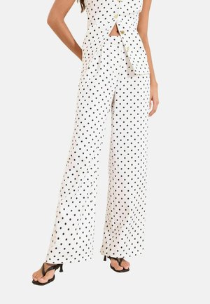 Kvinde iført hvide bukser med vide ben og ærmeløs vest med sorte polkaprikker, kombineret med sorte sandaler med hæl.