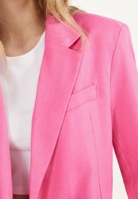 Blazer rosa hecho de tejido texturizado, con un diseño ajustado, solapas con muesca y un bolsillo en el pecho. Acompañado de una blusa blanca de canalé.