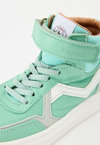 Zapatillas altas en cuero verde menta con acentos grises. Cuentan con cordones, una correa de velcro y una etiqueta con el logo en la lengua. Textura suave.