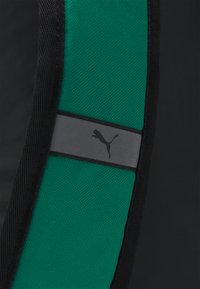 Un tissu athlétique noir présente une bande texturée verte, bordée de maille noire. Un bandeau gris affiche un logo Puma noir.
