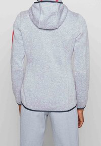 Grijze hoodie van een gestructureerde stof, met een verstelbare capuchon en contrasterende afwerking. Rode accenten op de mouwen en een aansluitende snit.