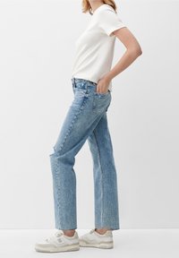 Mujer con jeans rectos de color azul claro, camiseta blanca de manga corta y zapatillas blancas, de pie de lado con una mano en el bolsillo.