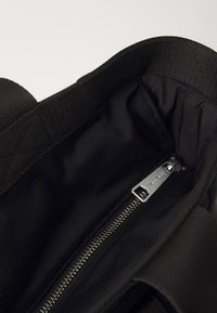 Intérieur d'un sac fourre-tout en tissu noir, doté d'une texture lisse, d'une poche zippée et d'un tirette de fermeture éclair en argent de marque avec un revêtement foncé.