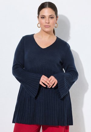 Kvinde med mørkt hår iført navyblå ribstrikket sweater med udsvungne ærmer og røde bukser, stående mod en hvid baggrund.