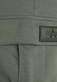 Grüner Stoff mit gerippter Textur, versehen mit einem gestickten Design und einem schwarzen Patch, auf dem "ALPHA INDUSTRIES" und Logodetails eingeprägt sind.
