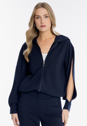 Blondynka z luźnymi falami włosów ma na sobie granatową bluzę na suwak z otwartymi, rozciętymi rękawami, pod którą znajduje się biała koszulka na ramiączkach, oraz granatowe spodnie, stojąca na tle białego tła.